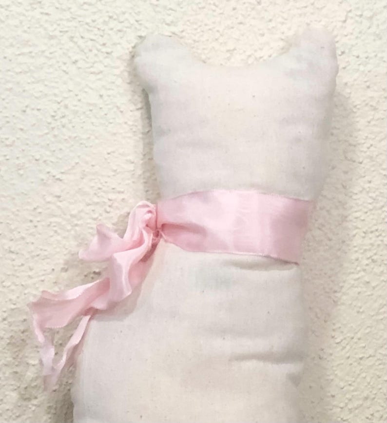 May include: A white cat-shaped stuffed toy with a light pink ribbon tied around its middle. The ribbon is knotted into a bow on the left side. The toy has a simple, minimalist design.
