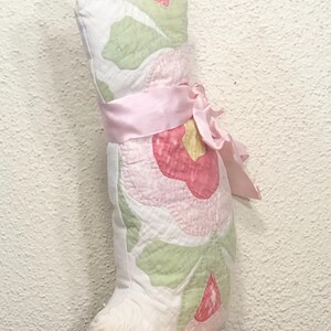 May include: A handmade cat-shaped pillow with a floral pattern in pink, green, and white. The pillow is tied with a pink ribbon and bow. The pillow is made of quilted fabric and has a soft, plush appearance.
