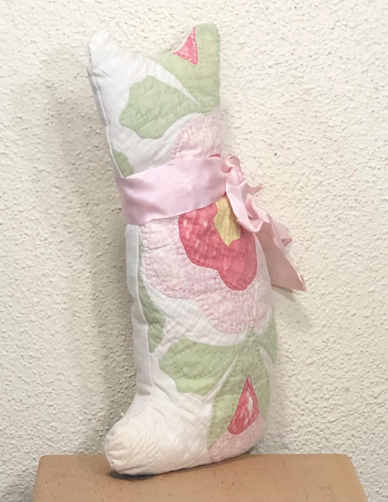 May include: A handmade cat-shaped pillow with a floral pattern in pink, green, and white. The pillow is tied with a pink ribbon and bow. The pillow is made of quilted fabric and has a soft, plush appearance.