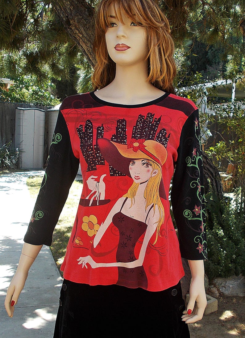 May include: A red, black, and green long-sleeved shirt with a graphic print of a woman in a hat and dress holding a shopping bag. The graphic print includes a cityscape in the background.