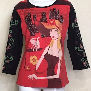 May include: Red long-sleeved shirt with a black and green floral design on the sleeves. The front of the shirt features a graphic of a woman in a hat holding a shopping bag with a city skyline in the background.