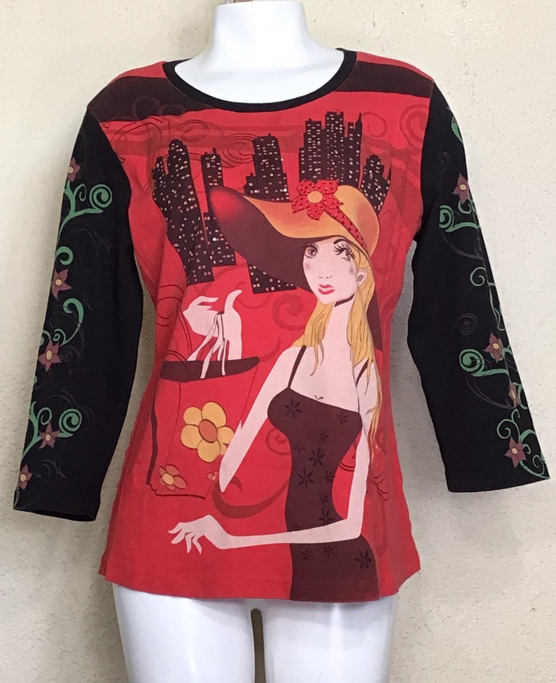 May include: Red long-sleeved shirt with a black and green floral design on the sleeves. The front of the shirt features a graphic of a woman in a hat holding a shopping bag with a city skyline in the background.