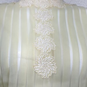 May include: Pale yellow blouse with vertical stripes and white floral lace embellishments. The high neckline and center front feature delicate, embroidered floral appliques. The sheer fabric creates a light and airy look.