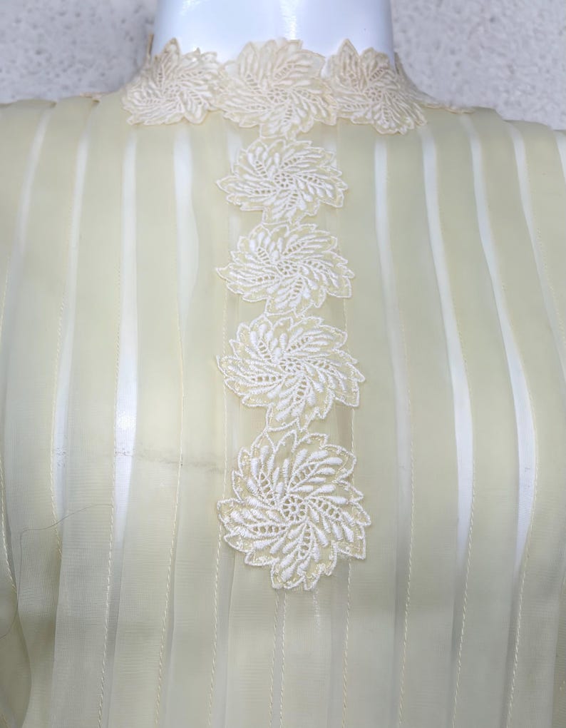 May include: Pale yellow blouse with vertical stripes and white floral lace embellishments. The high neckline and center front feature delicate, embroidered floral appliques. The sheer fabric creates a light and airy look.