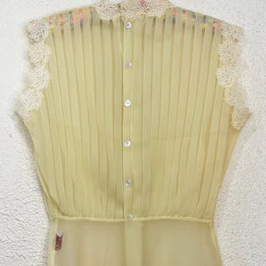 May include: A pale yellow sleeveless blouse with vertical pleats and a high neckline. The blouse features delicate floral lace trim along the neckline and armholes. It has a button-down front with small, round buttons.