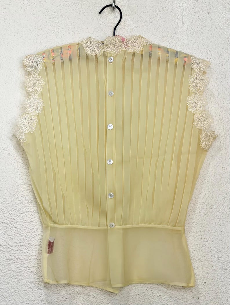 May include: A pale yellow sleeveless blouse with vertical pleats and a high neckline. The blouse features delicate floral lace trim along the neckline and armholes. It has a button-down front with small, round buttons.