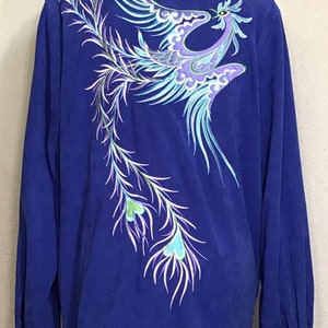 May include: A royal blue long-sleeved shirt with a colorful embroidered phoenix design on the back. The phoenix is depicted in flight with its wings spread wide and its tail feathers trailing behind it.