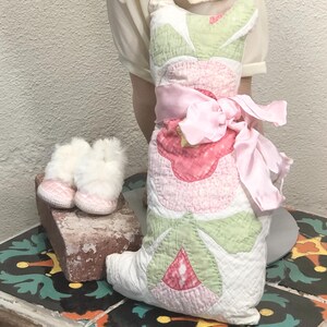 May include: A quilted cat-shaped pillow with pink and green floral designs, tied with a pink ribbon. Beside it are small white and pink baby booties. The items are displayed on a decorative tile surface.