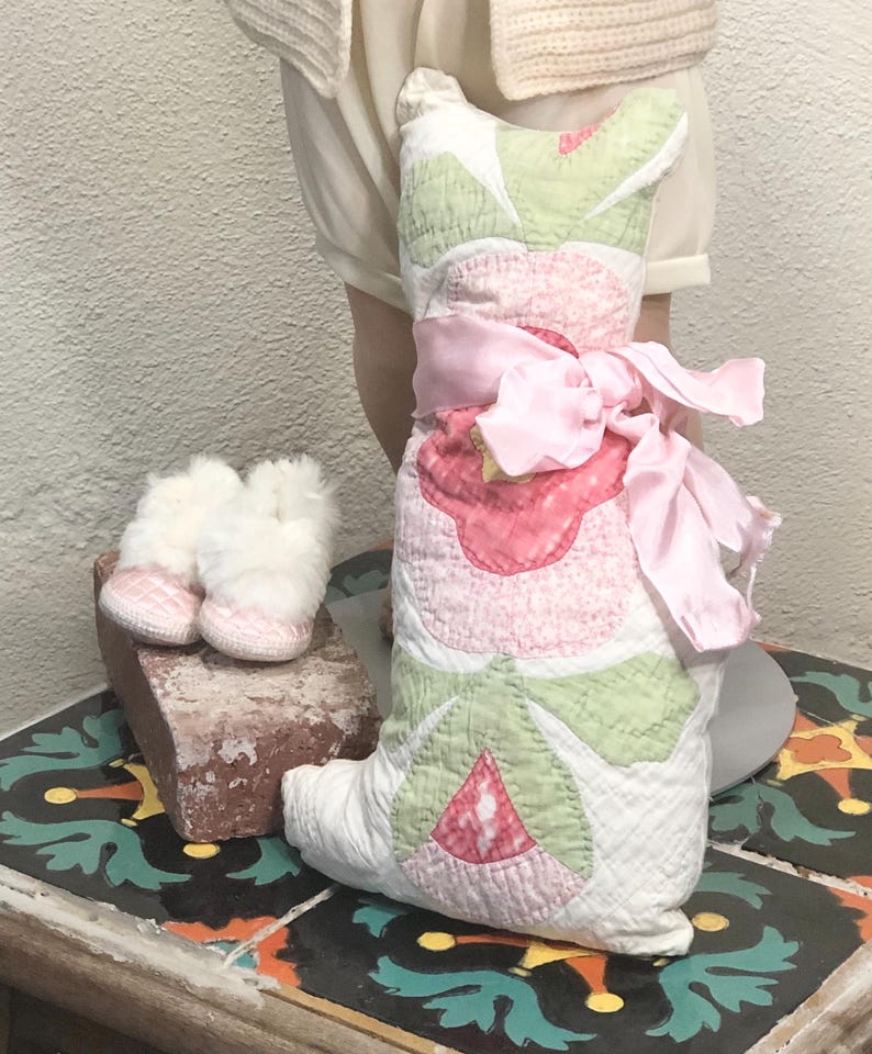 May include: A quilted cat-shaped pillow with pink and green floral designs, tied with a pink ribbon. Beside it are small white and pink baby booties. The items are displayed on a decorative tile surface.