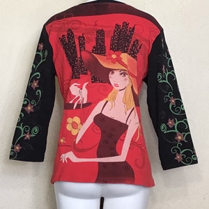 May include: Red long-sleeved shirt with a black and green floral design on the sleeves. The front of the shirt features a graphic of a woman in a red hat and a black dress, holding a shopping bag in front of a city skyline.