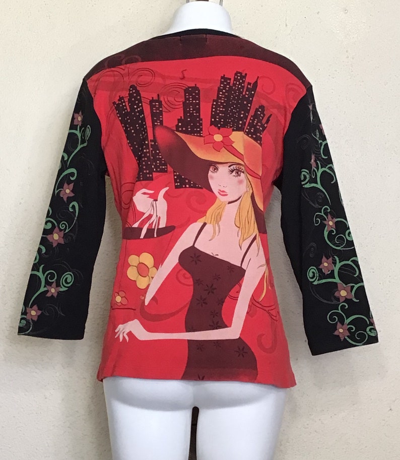 May include: Red long-sleeved shirt with a black and green floral design on the sleeves. The front of the shirt features a graphic of a woman in a red hat and a black dress, holding a shopping bag in front of a city skyline.