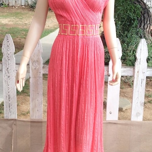 May include: A coral-coloured sleeveless dress with a V-neckline and a gold geometric pattern on the waist. The dress has a pleated texture and falls to the mid-calf. A matching red necklace is worn.
