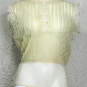 May include: A pale yellow vintage blouse with a high neckline and short sleeves. The blouse features vertical pleats and delicate floral lace trim around the neckline, shoulders, and down the front. The bottom portion is a solid cream colour.