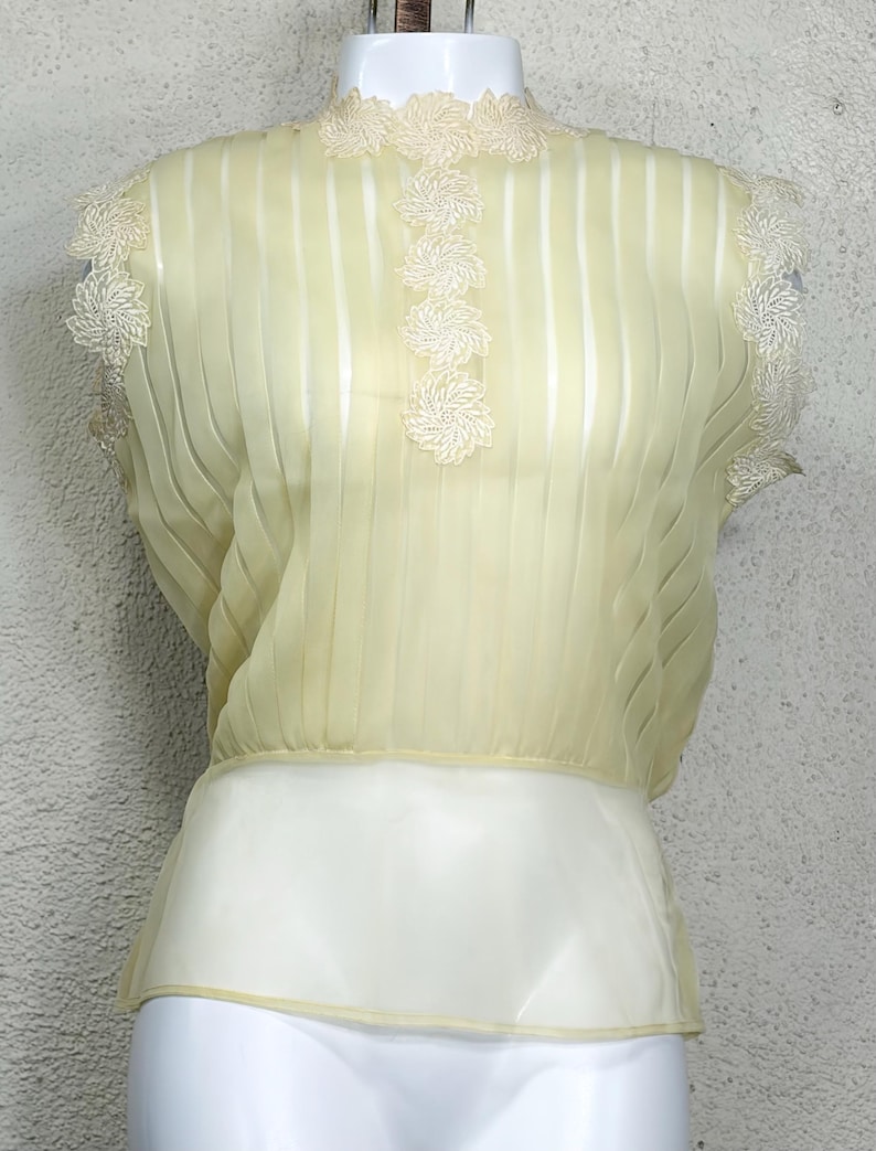 May include: A pale yellow vintage blouse with a high neckline and short sleeves. The blouse features vertical pleats and delicate floral lace trim around the neckline, shoulders, and down the front. The bottom portion is a solid cream colour.