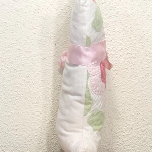 May include: A decorative, handmade pillow in a teardrop shape. The pillow features a white fabric with pink and green floral patterns. A pink satin ribbon is tied around the middle, with a bow on the side. The pillow is resting on a wooden surface.
