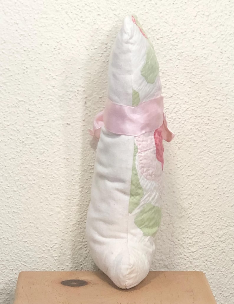 May include: A decorative, handmade pillow in a teardrop shape. The pillow features a white fabric with pink and green floral patterns. A pink satin ribbon is tied around the middle, with a bow on the side. The pillow is resting on a wooden surface.