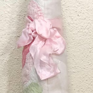 May include: A soft, light pink fabric item with a large, ruffled bow. The item has a patchwork design with shades of pink and white. The bow is made of a silky material, adding a delicate touch to the overall design.