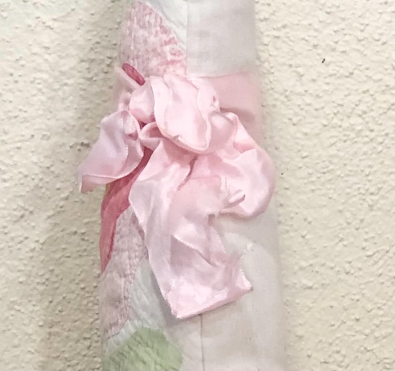 May include: A soft, light pink fabric item with a large, ruffled bow. The item has a patchwork design with shades of pink and white. The bow is made of a silky material, adding a delicate touch to the overall design.