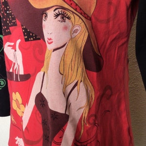 May include: Red and black long-sleeved shirt with a graphic print of a woman wearing a yellow hat with a red flower and rhinestones. The woman is holding a red bag with a yellow flower.