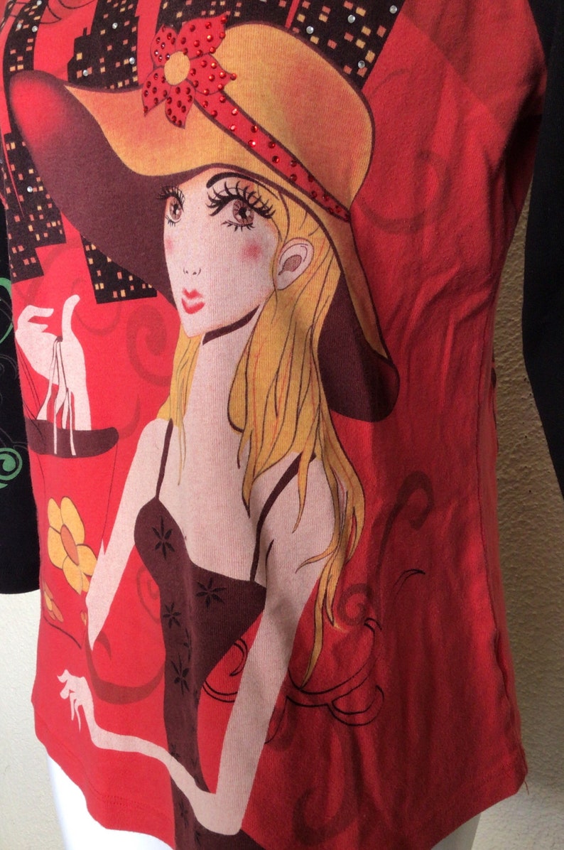 May include: Red and black long-sleeved shirt with a graphic print of a woman wearing a yellow hat with a red flower and rhinestones. The woman is holding a red bag with a yellow flower.