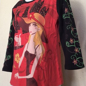 May include: Red, black, and green long-sleeved shirt with a graphic print of a woman wearing a hat and holding a scale. The woman is standing in front of a cityscape.
