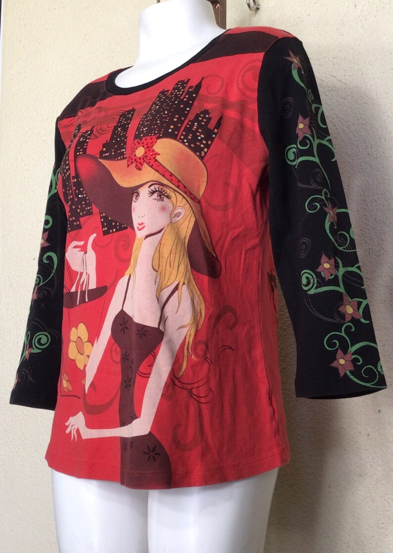 May include: Red, black, and green long-sleeved shirt with a graphic print of a woman wearing a hat and holding a scale. The woman is standing in front of a cityscape.