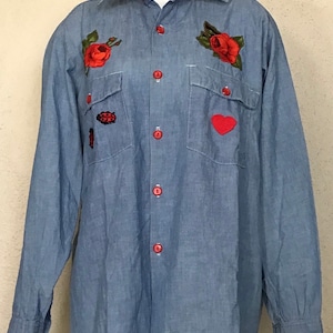 May include: A light blue denim shirt with red embroidered roses, a ladybug, and a heart. The shirt has a button-up front and long sleeves.