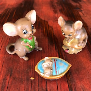 May include: Three ceramic mouse figurines. One mouse is standing and wearing a green vest. Another mouse is sitting on a leaf. The third figurine shows two mice in a boat. The figurines are brown, with white and black accents.