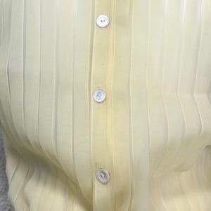 May include: A close-up of a pale yellow, sheer button-down shirt. The shirt has vertical stripes and small, white, round buttons down the centre. The fabric appears lightweight and slightly translucent.