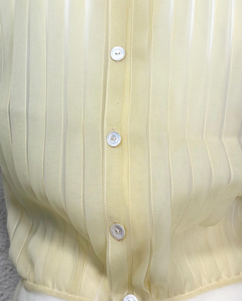 May include: A close-up of a pale yellow, sheer button-down shirt. The shirt has vertical stripes and small, white, round buttons down the centre. The fabric appears lightweight and slightly translucent.