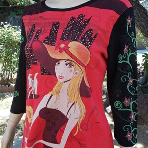 May include: Red, black, and green long-sleeved shirt with a graphic print of a woman wearing a large hat and a black dress. The background features a cityscape.