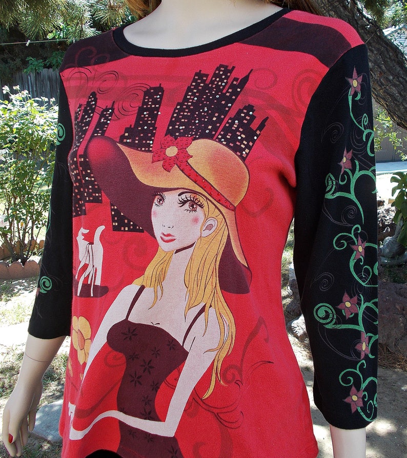 May include: Red, black, and green long-sleeved shirt with a graphic print of a woman wearing a large hat and a black dress. The background features a cityscape.