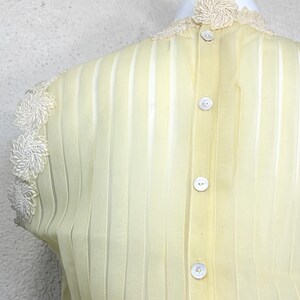 May include: Pale yellow blouse with vertical stripes and a high, lace-trimmed collar. The back of the blouse features a row of white buttons. Delicate white lace adorns the shoulders, adding a vintage touch to the garment.