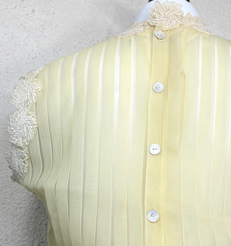 May include: Pale yellow blouse with vertical stripes and a high, lace-trimmed collar. The back of the blouse features a row of white buttons. Delicate white lace adorns the shoulders, adding a vintage touch to the garment.