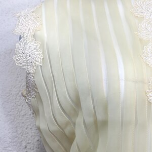 May include: Close-up of a cream-coloured garment with vertical striped detailing. Delicate white floral lace trim adorns the shoulder and neckline. The fabric appears sheer and lightweight, suggesting a formal or vintage style.