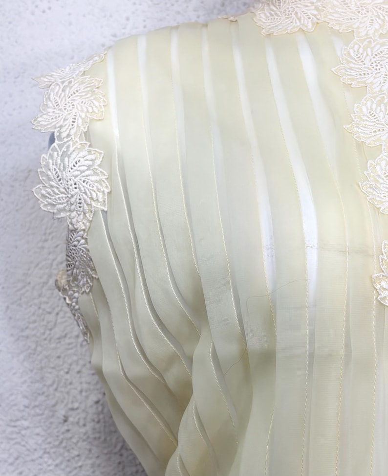 May include: Close-up of a cream-coloured garment with vertical striped detailing. Delicate white floral lace trim adorns the shoulder and neckline. The fabric appears sheer and lightweight, suggesting a formal or vintage style.
