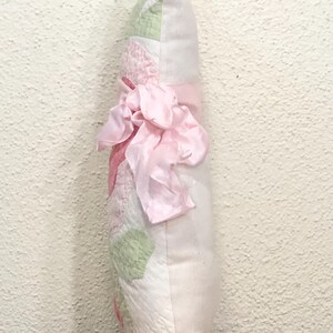 May include: A decorative pillow with a patchwork design in white, pink, and green. A large pink ribbon is tied around the center. The pillow is approximately 12 inches long.
