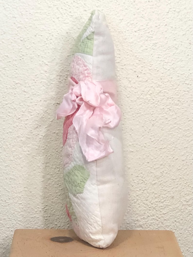May include: A decorative pillow with a patchwork design in white, pink, and green. A large pink ribbon is tied around the center. The pillow is approximately 12 inches long.