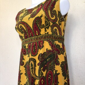 May include: A sleeveless maxi dress with a yellow background and a paisley pattern in shades of red, green, and brown. The dress has a narrow band of gold trim around the waist.