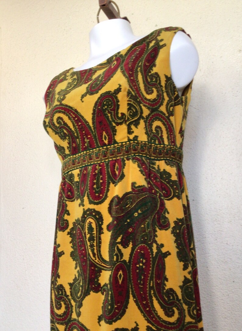 May include: A sleeveless maxi dress with a yellow background and a paisley pattern in shades of red, green, and brown. The dress has a narrow band of gold trim around the waist.