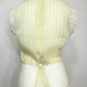 May include: Pale yellow vintage blouse with vertical pleats and a row of white buttons down the back. The blouse features delicate lace detailing on the collar and sleeves, and a sheer, flowing fabric.