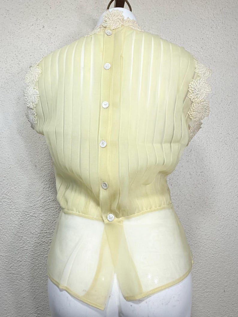 May include: Pale yellow vintage blouse with vertical pleats and a row of white buttons down the back. The blouse features delicate lace detailing on the collar and sleeves, and a sheer, flowing fabric.