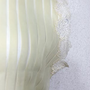 May include: Close-up of a cream-coloured garment with vertical, wavy, sheer stripes. The edge features delicate white lace floral detailing. The fabric appears soft and flowing, suggesting a dress or top.