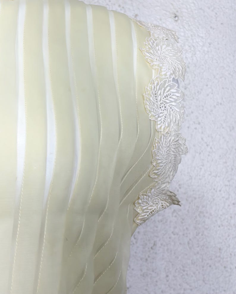 May include: Close-up of a cream-coloured garment with vertical, wavy, sheer stripes. The edge features delicate white lace floral detailing. The fabric appears soft and flowing, suggesting a dress or top.