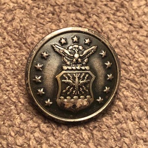 May include: A round, silver-toned button with an embossed design. The button features an eagle with outstretched wings above a shield, surrounded by stars. The button has a slightly aged appearance.
