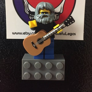 May include: A gray Lego figurine with a gray beard and sunglasses playing a brown acoustic guitar. The figurine is standing on a gray Lego base with the word "LEGO" printed on each block.