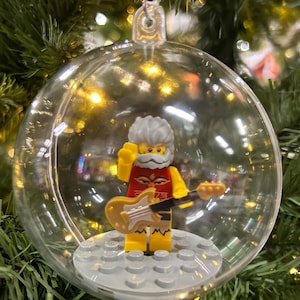 May include: A clear, round Christmas ornament with a Lego figure inside. The Lego figure has a white beard, gray hair, and is wearing a red and yellow outfit. It is holding a yellow guitar. The ornament is hanging from a Christmas tree.