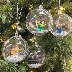 May include: Clear Christmas ornaments with Lego figures inside, hanging on a green Christmas tree. The ornaments contain Lego figures playing musical instruments. The ornaments are round and transparent, with silver string hangers.