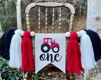 May include: A red tractor with the word "one" printed on a burlap banner with black and white, and red yarn tassels.