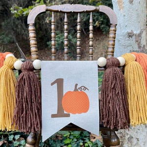 May include: A brown wooden chair with a banner that says "1" with a pumpkin on it. The banner is decorated with brown, orange, and yellow yarn tassels.